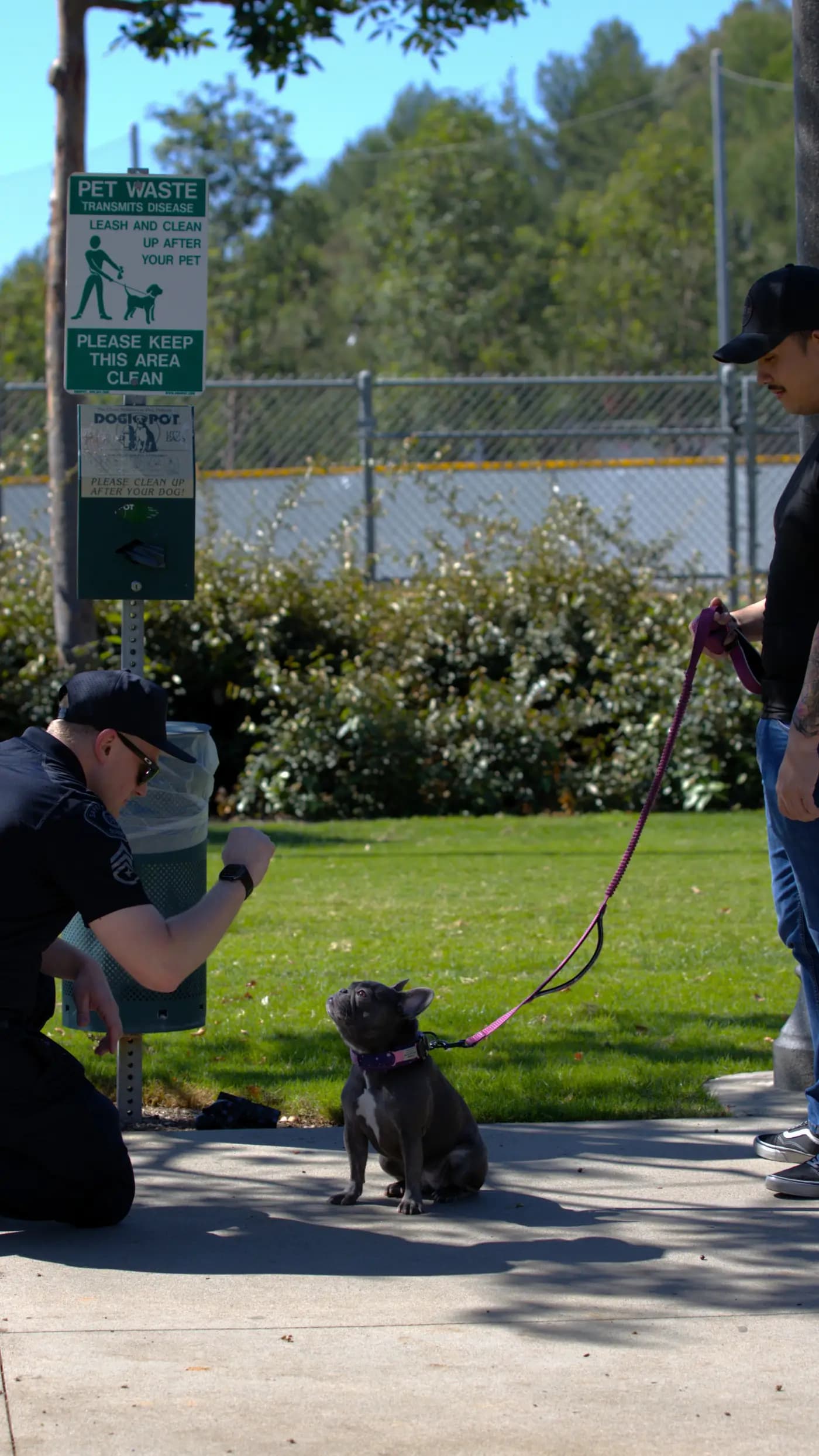 Officer greeting a resident's dog beside a pet-waste station in a park