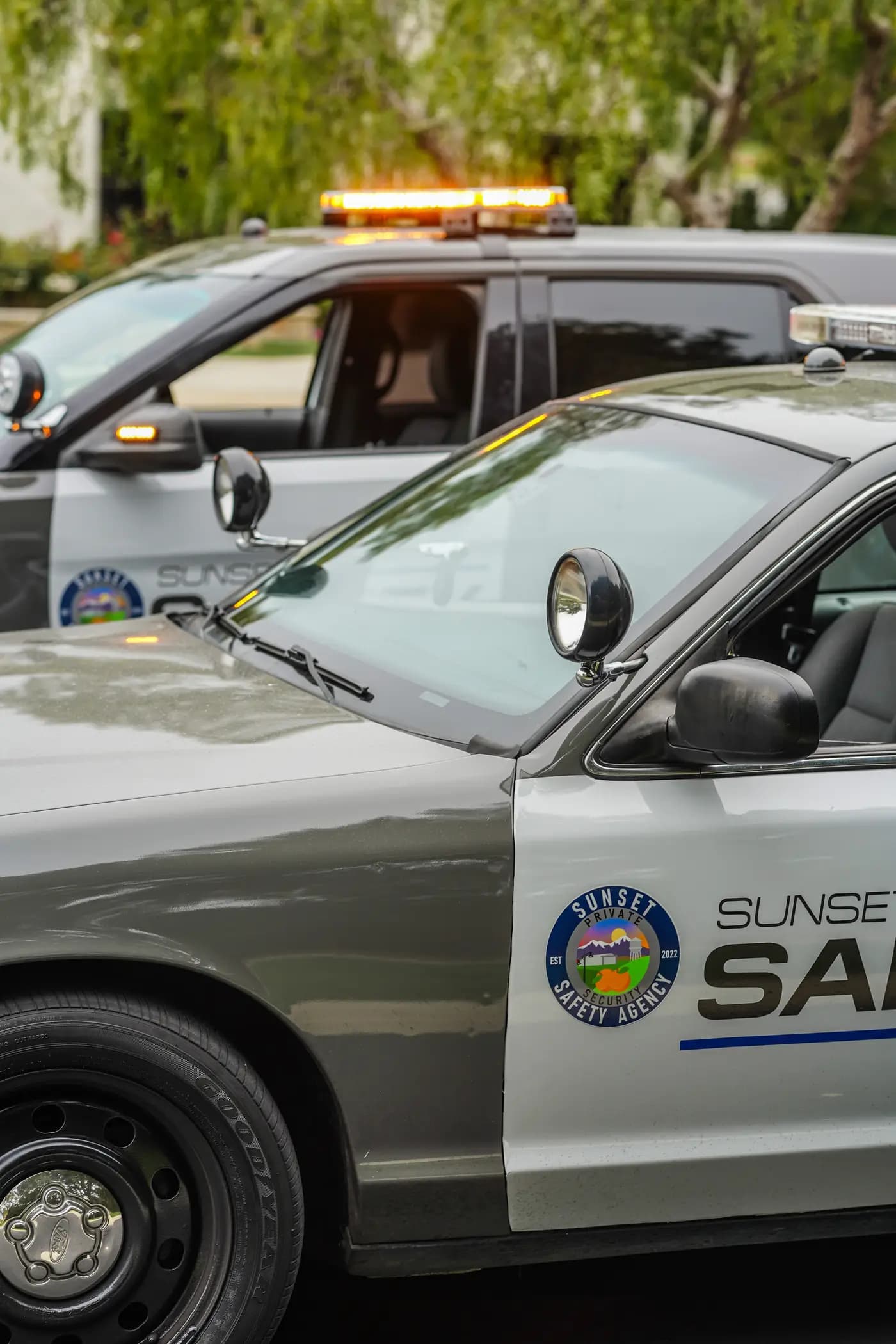 Two patrol cars staged nose-to-tail with amber lightbars lit on a tree-lined street