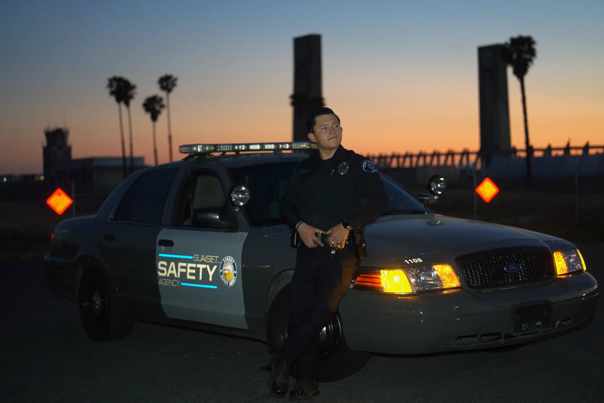 Officer at the fender of a patrol unit at twilight with palms silhouetted on the orange horizon