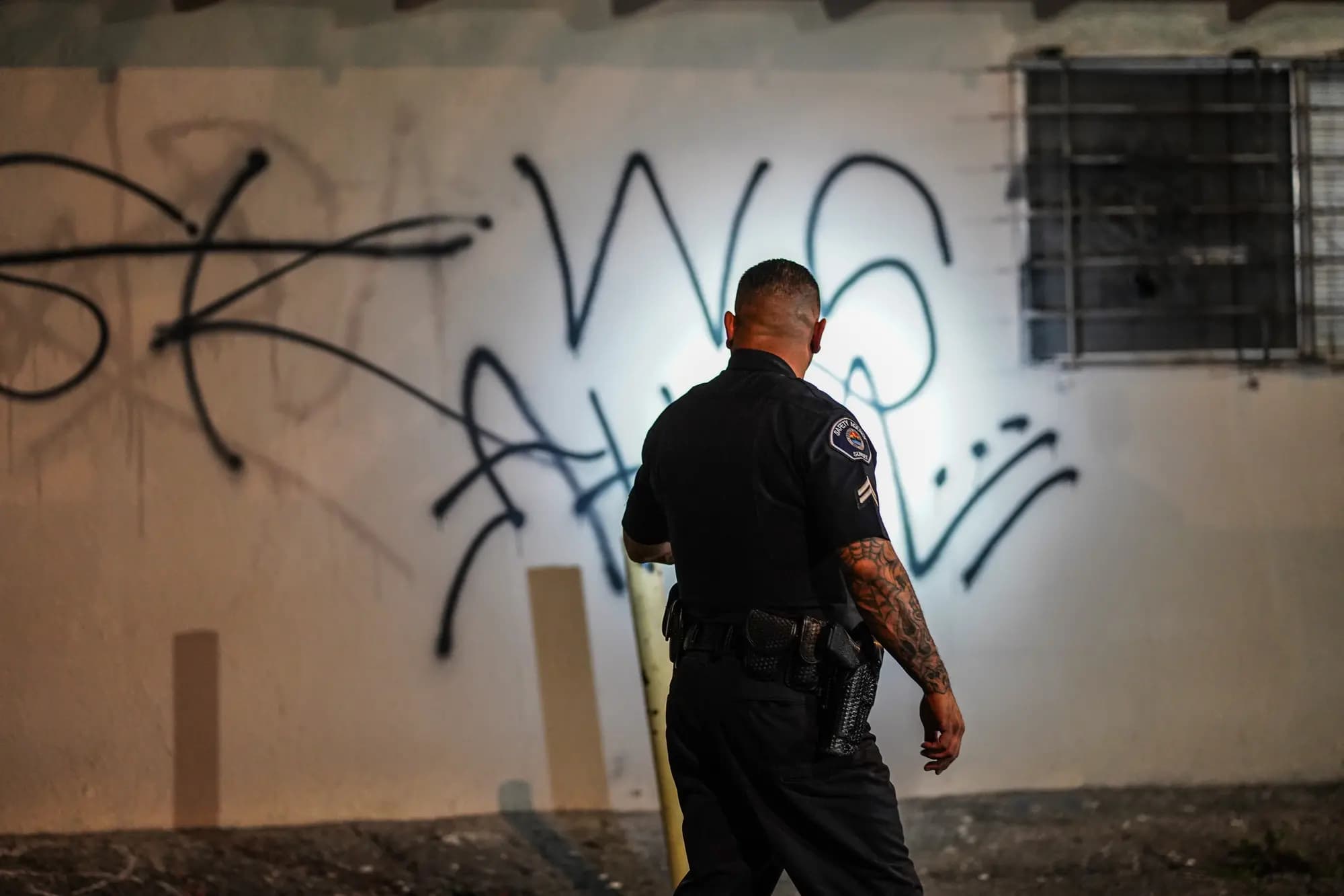 Officer's flashlight beam lighting up graffiti tags on an apartment wall during a night patrol