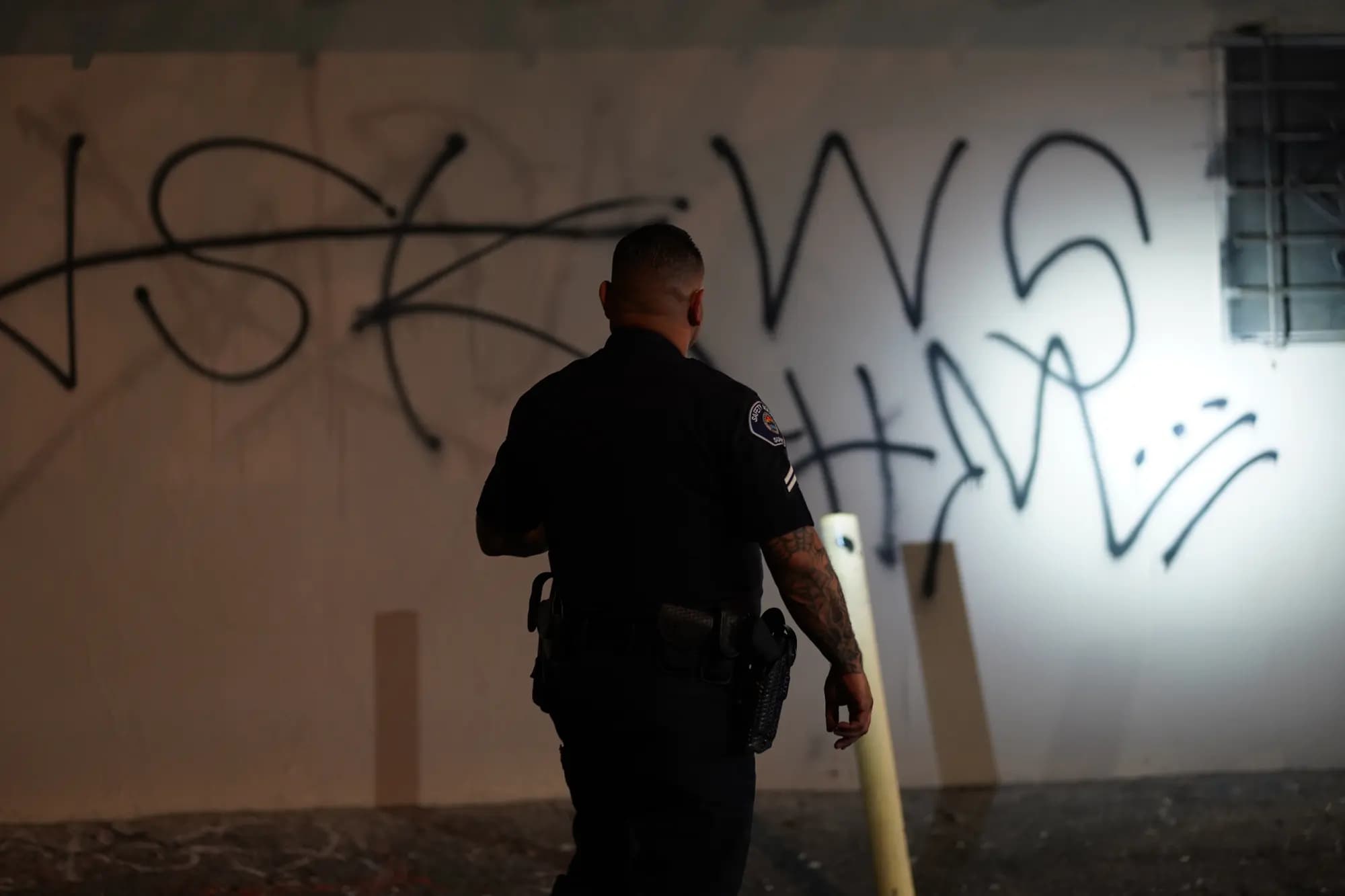 Silhouetted officer documenting a floodlit graffiti wall on a night vandalism check