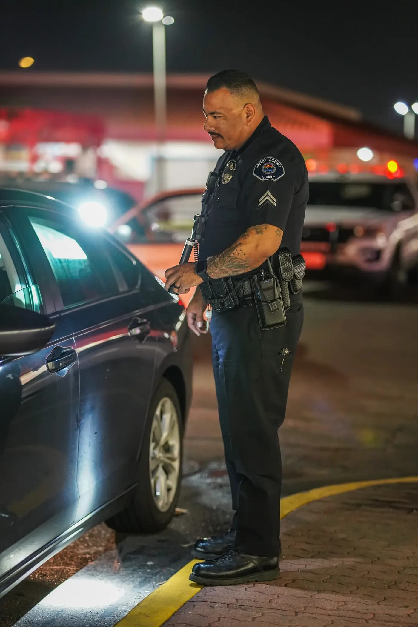 Corporal inspecting a sedan with a flashlight in a retail parking lot at night
