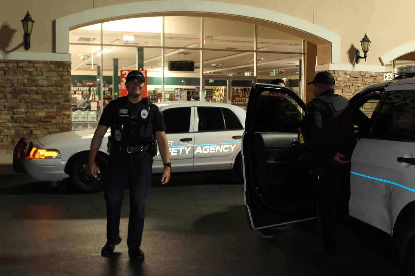 Officer walking a retail storefront at night with patrol cars behind