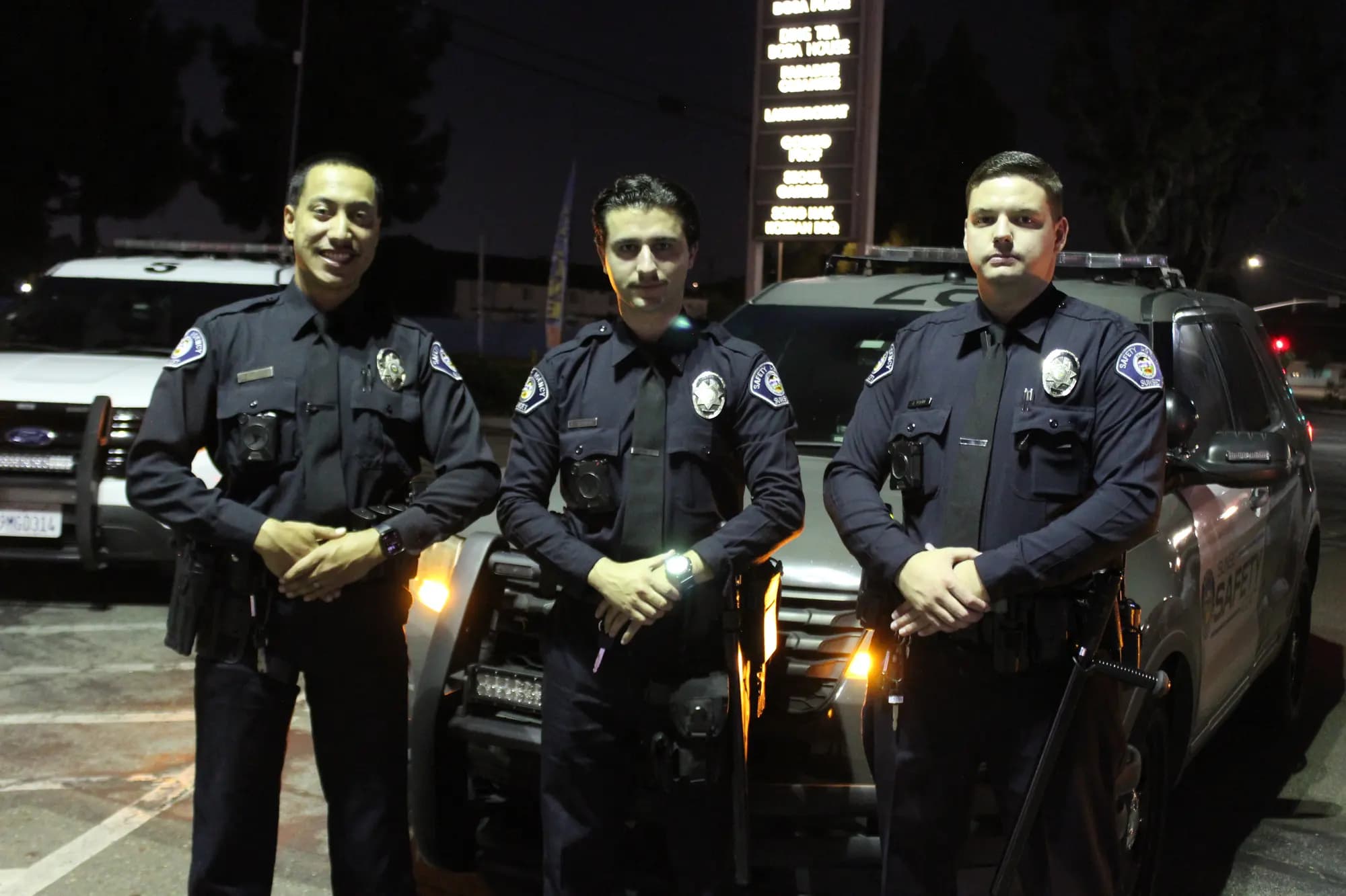 Three officers in Class A uniforms with body cameras standing shoulder to shoulder at night