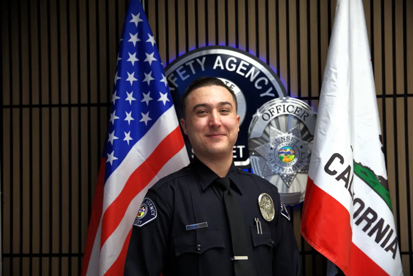 Officer smiling in uniform and tie between the US and California flags