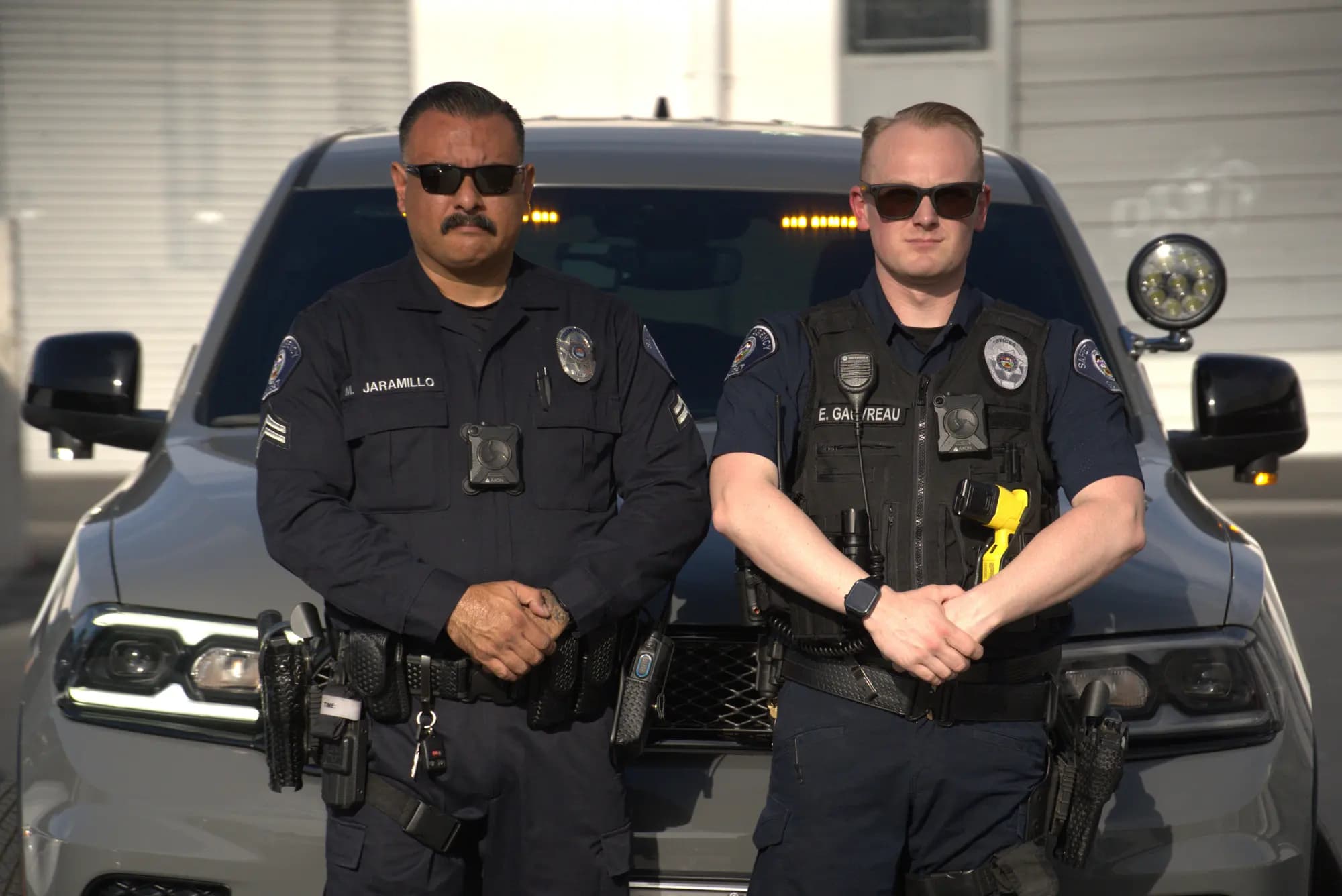 Two officers pose arms-crossed in front of a gray patrol SUV in daylight